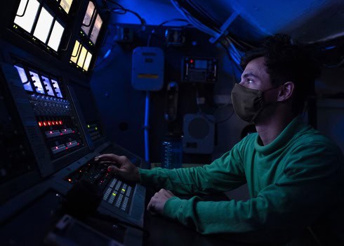 Control room operator monitoring multiple screens and equipment in a dark, high-tech environment.